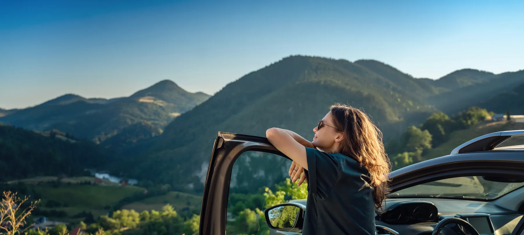 A woman resting her arms on an open car door, looking out over a mountain valley at sunset
